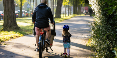 Ein Mann fährt auf einem Fahrrad auf einem schönen Radweg. Daneben ist ein Kind auf einem kleinen Roller.